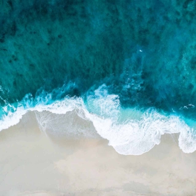 Aerial view wallpaper featuring turquoise waves crashing onto a sandy beach. Foamy white surf contrasts with light beige sand. Varying shades of blue highlight the water's depth and movement, creating a stunning natural scene from above. Keywords: Turquoise waves, foamy surf, beige sand, varying blue shades, aerial beach view.