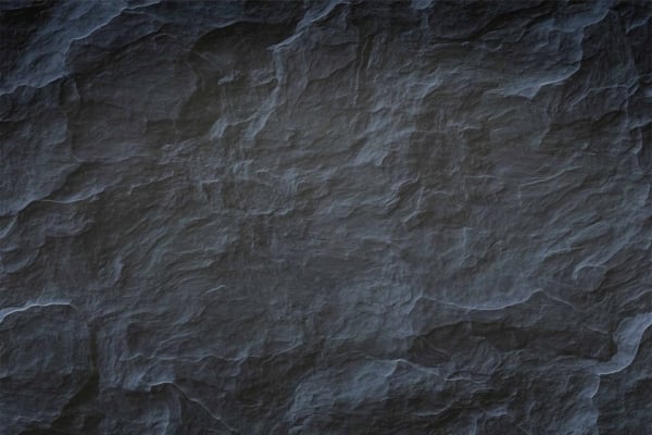 A close-up view of a dark, rough, and textured rock surface with thin white veins running across it. The weathered rock displays varying shades of gray with subtle highlights, creating a rugged pattern reminiscent of Black Widow's resilience. Elements: dark rock texture, white veins, varying gray shades.