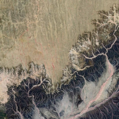 A satellite view wallpaper of rugged desert terrain with intricate erosion patterns. It features light and dark hues, winding riverbeds, and sections of visible mountains.