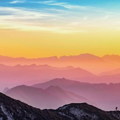 Stunning landscape wallpaper: Silhouetted hiker on mountain ridge, overlooking hazy mountain ranges. Vibrant sunset with hues of orange, pink, and purple. Clear sky transitioning into twilight. Elements: silhouetted hiker, mountain ridge, vibrant sunset colors (orange, pink, purple), hazy mountain ranges.