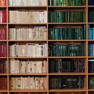 A library bookshelf background featuring a gradient of color-organized books. The transition starts with red books on the left, moving through beige and green shades, and concludes with blue and black books on the right. This wallpaper emphasizes elements such as the vibrant mix of book spines, creating a colorful spectrum.