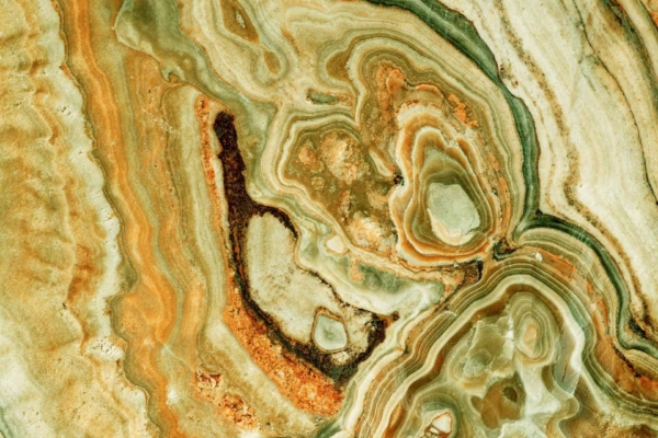 A close-up of a polished stone surface reveals intricate patterns in shades of green, brown, and beige. The swirling designs and rich textures highlight the natural beauty and complexity of minerals formed beneath the Earth.