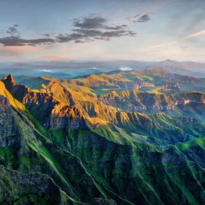A breathtaking aerial view of the Drakensburg mountain range during sunset. Peaks highlighted with golden sunlight, casting shadows over lush green valleys below. Distant mountains and a sky with scattered clouds add drama and serenity. Elements: peaks, golden sunlight, shadows, green valleys, distant mountains, scattered clouds. Colors: gold, green, blue sky with white clouds.