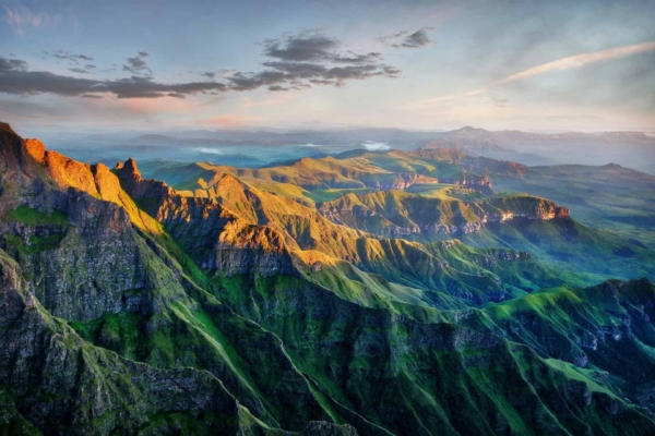 A breathtaking aerial view of the Drakensburg mountain range during sunset. Peaks highlighted with golden sunlight, casting shadows over lush green valleys below. Distant mountains and a sky with scattered clouds add drama and serenity. Elements: peaks, golden sunlight, shadows, green valleys, distant mountains, scattered clouds. Colors: gold, green, blue sky with white clouds.