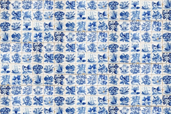 Blue and white ceramic tile wallpaper with intricate patterns including flowers, sailing boats, birds, and other motifs. Hand-painted designs evoke Dutch heritage. Delicate and detailed artistry in a uniform grid.