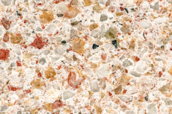 A close-up of a terrazzo surface features a coarse, speckled texture with multi-colored chips of red, brown, white, and green. These chips vary in shape and size, set in a light-colored base. The wallpaper highlights the dynamic mix of colors and irregular patterns that define terrazzo's unique aesthetic.