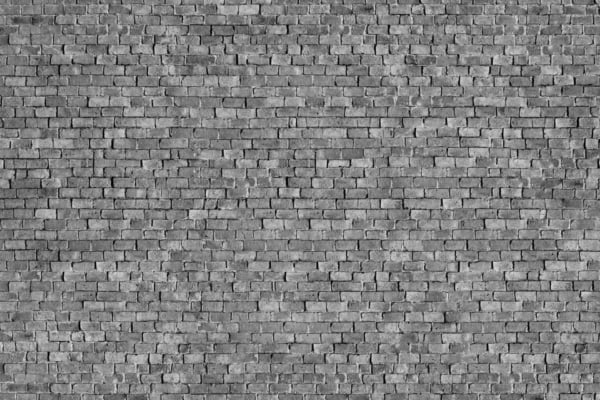 A close-up of a weathered gray brick wall with evenly stacked rectangular bricks. The surface has minor wear and irregularities, giving a rough but uniform appearance. Colors include various shades of gray, with slight discolorations adding texture. No signs or graffiti are present.