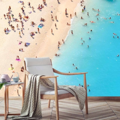 An overhead beach view wallpaper with a crowded shoreline. Dominant colors: sand beige, turquoise water, and clusters of people. Foreground features: cozy armchair with blanket, small side table with a teapot and cup on wooden floorboards.
