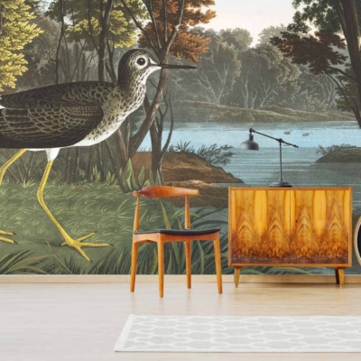 The living room features a large, nature-themed wall mural depicting a wading bird by the riverbank in a forest. The wallpaper includes elements such as lush greenery, flowing water, trees, and the detailed figure of a bird. Colors range from various shades of green and brown to blue highlights of the river and subtle touches of white on the bird's feathers.
