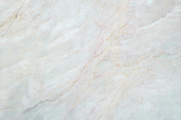 A close-up view of a light-colored marble surface with subtle veining in shades of beige, cream, and soft pink. The texture is smooth, with natural patterns creating an elegant, polished appearance. Highlights include delicate lines in beige, cream, and soft pink tones.
