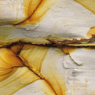 Close-up image of a rock surface displaying intricate layers and patterns. Swirling bands of orange, yellow, and white create an abstract design. Rough texture with visible striations and cracks.