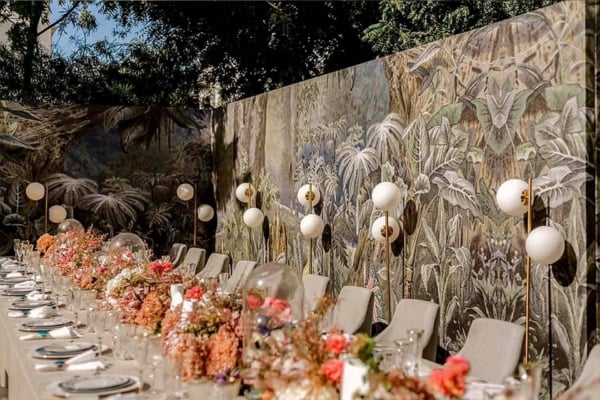 The wallpaper features a lush, jungle-themed mural with rich shades of green, interspersed with tropical foliage and vibrant flowers in hues of red, orange, and yellow. Exotic animals and birds are subtly included within the dense vegetation. The background displays varying depths of green to create a sense of depth and realism, while darker shadows add contrast.