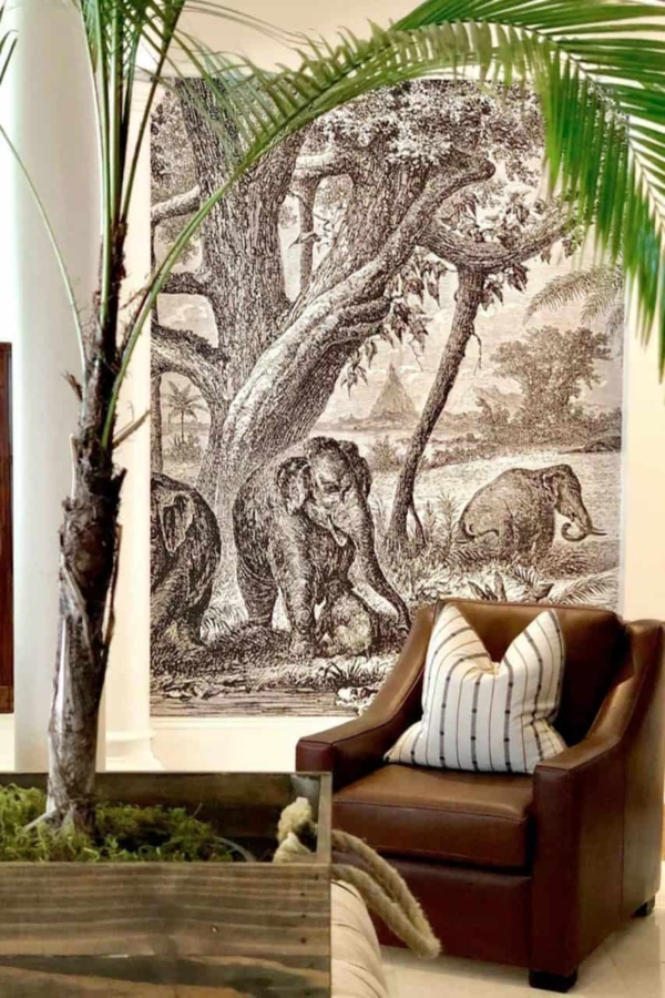 The wallpaper features a large, detailed mural of elephants in a natural landscape. It showcases rich earth tones, with shades of grey and brown for the elephants, lush greens for the foliage, and soft blues for the sky. This naturalistic artwork brings depth and a touch of wilderness to any interior space.