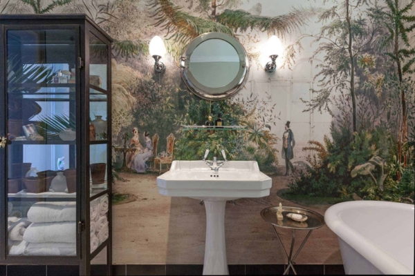 The wallpaper is a mural depicting a lush, nature-inspired scene featuring vibrant greens, deep blues, and earthy browns. It showcases detailed foliage, with shades of emerald and moss green, interspersed with blooming flowers in hues of red, yellow, and pink. Birds in striking colors like blue and red are perched among the leaves. The overall aesthetic is reminiscent of a dense forest or tropical paradise.