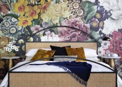 A stylish bedroom features a large floral mural behind the bed. The wallpaper highlights elements like roses, peonies, and lilies in shades of mustard yellow, dark blue, black, and white.