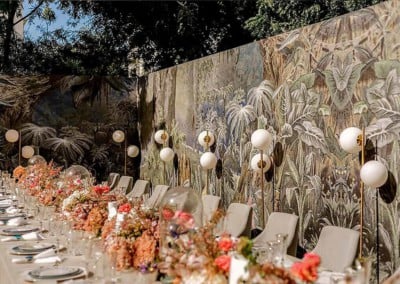 The wallpaper showcases lush, tropical foliage in vibrant greens and delicate leaf patterns. It features an array of botanical elements, including palm fronds and exotic plants. The design exudes a lively yet soothing atmosphere with prominent green shades, interspersed with hints of natural earth tones and a textured, layered appearance to mimic the depth of a tropical jungle.