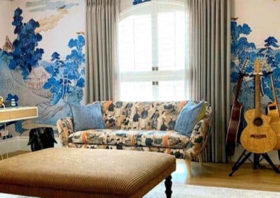 The wallpaper in the brightly lit living room showcases an intricate mural with a mix of vibrant and earthy tones. It features bold splotches of deep blue, hints of emerald green, and accents of warm golden hues. Delicate lines weave through abstract floral patterns, adding an artistic touch to the space.