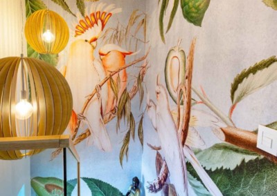 The mural wallpaper features vibrant, colorful birds and lush green foliage. The birds are depicted in a variety of bright hues—red, blue, yellow, and orange—contrasted against deep emerald leaves and delicate floral accents. Vivid details bring the scene to life with a rich spectrum of colors providing an eye-catching backdrop.