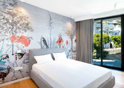 Introducing a modern bedroom that exudes tranquility and sophistication, featuring a stunning colorful botanical mural with birds on the wall behind the bed. The vibrant wallpaper showcases hues of green from lush foliage, bursts of yellow and red flowers, and intricate details of various bird species. These elements create a captivating visual centerpiece that beautifully contrasts with the room's minimalist design. Flooded with natural light from floor-to-ceiling windows adorned with gray curtains, this serene space offers an inviting ambiance perfect for relaxation. Ideal for any home decor portfolio, this room seamlessly integrates nature-inspired elegance into contemporary living spaces.
