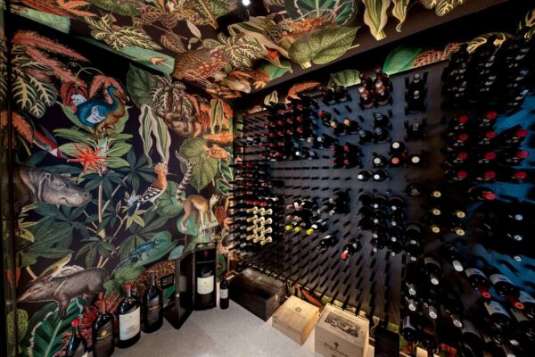 A wine cellar with walls adorned in vibrant, tropical-themed wallpaper featuring an array of exotic animals and colorful plants. The wallpaper bursts with hues of lush green palm leaves, bright pink flamingos, vivid toucans in black and yellow, brilliant blue macaws, and a splash of red hibiscus flowers. Richly detailed vines and foliage weave through the design, adding layers of emerald shades interspersed with accents of orange butterflies and purple orchids. This dynamic backdrop exudes a lively jungle atmosphere that captures attention instantly.