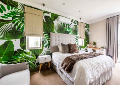 The wallpaper features a tropical theme with large, vibrant green leaves that create an immersive, lush atmosphere. The pattern includes various shades of green and subtle accents of earthy tones to mimic natural foliage. The design is bold yet elegant, providing a striking backdrop for the bedroom's decor.