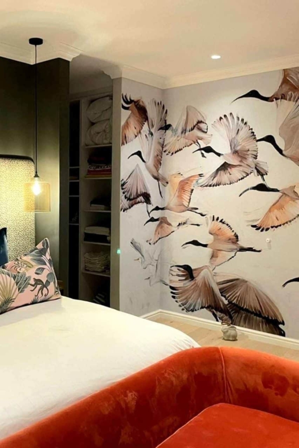 A cozy bedroom showcases a mural of flying birds on the wall. The wallpaper features shades of blue skies streaked with soft white clouds, depicting birds in various stages of flight. Feather details and subtle gradients add depth, while touches of green from tree branches integrate seamlessly into the design, highlighting natural elements and tranquil colors.