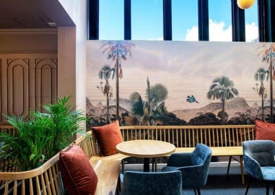 The wallpaper features a large mural depicting tropical scenery, showcasing lush greenery with vibrant leaves and colorful flowers. Hints of ocean blue, sunset hues, and earthy tones weave through the design. Natural elements like palm fronds and exotic birds add to its inviting charm. The overall palette combines rich greens, soft pastels, warm oranges and sunlit yellows – perfect for creating a cozy yet stylish atmosphere.