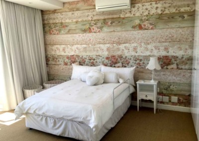 The floral-patterned wallpaper is the focal point, featuring a blend of soft pastels and bright hues. The design includes delicate pink roses, lush green leaves, and subtle hints of lavender. This charming pattern adds warmth and texture to the wooden wall, creating a cozy yet vibrant atmosphere perfect for a bedroom interior.