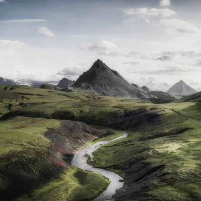 Expansive landscape wallpaper featuring a lone mountain peak under a cloudy sky. Gentle, winding river in the foreground cuts through lush green fields and rolling hills. Serene and picturesque scene with natural colors of greens, blues, and soft grays.
