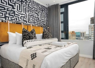 The wallpaper in the modern bedroom features a striking geometric pattern, primarily in black and white. The design comprises a series of interlocking shapes and lines that create a bold visual effect. This black-and-white geometric wallpaper serves as an eye-catching focal point behind the cushioned headboard.