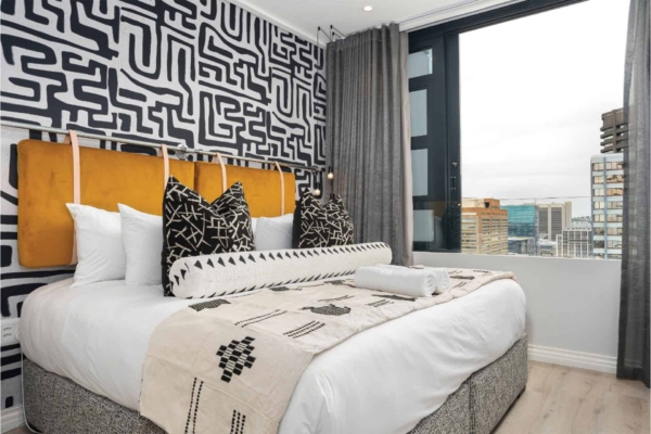 The wallpaper in the modern bedroom features a striking geometric pattern, primarily in black and white. The design comprises a series of interlocking shapes and lines that create a bold visual effect. This black-and-white geometric wallpaper serves as an eye-catching focal point behind the cushioned headboard.