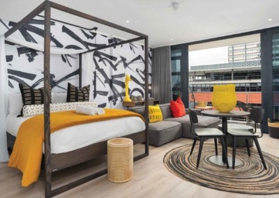 Modern hotel room wallpaper features a subtle geometric pattern in neutral tones, grey and white. Small accents of yellow lines mirror the room's yellow blanket and pillows, seamlessly tying the space together. The wallpaper intelligently integrates hints of red to complement the red pillows on the sectional sofa.