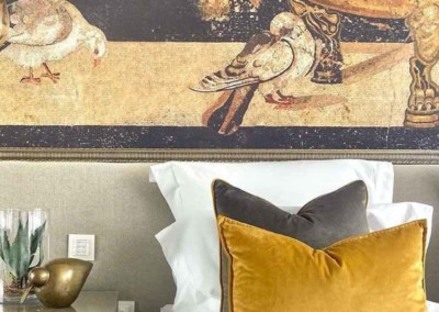 An elaborate mural features prominently on the wall above the bed, depicting birds and an urn. The wallpaper includes intricate elements such as detailed avian figures in flight and perched positions, alongside an ornate urn adorned with floral motifs. The color palette is rich and varied with hues of muted gold, soft grays, delicate whites, dusty blues, and touches of earthy greens. This elegant design adds a sophisticated focal point to the room’s decor.