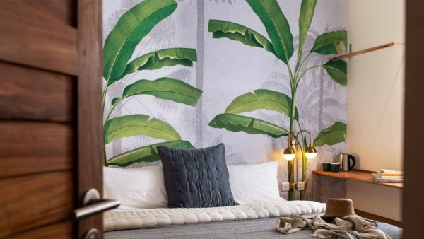 The wallpaper behind the bed features a large tropical leaf mural with lush, green foliage. The leaves are intricately detailed, depicting various shades of green from deep emerald to lighter lime. Hints of shadow create depth, emphasizing the natural texture and layers of the leaves.