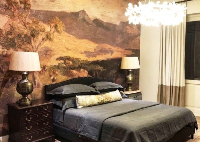 The wallpaper features a mural of a mountainous landscape with stunning detail. Dominant elements include towering, snow-capped peaks, lush green forests at the base, and a serene blue sky with soft white clouds. The colors range from rich forest greens and deep blues to lighter hues of grey and white for the mountains and clouds. Perfectly textured to add depth and tranquility to any space, this nature-inspired mural creates a breathtaking focal point in the bedroom design.