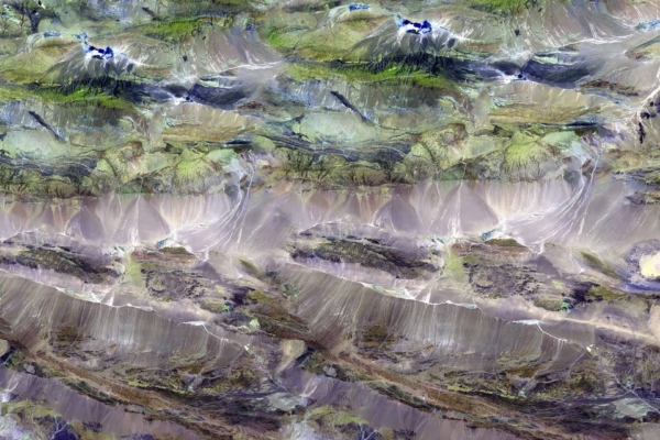 Aerial view wallpaper of a rugged, mountainous landscape. Featuring a mix of green and brown hues, the terrain resembles Peri Peri spice with deep valleys, scattered vegetation patches, and shadowed areas indicating elevation changes. Complex textures highlight natural formations such as rocky cliffs and ridges.