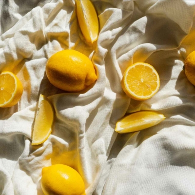 Lemon-themed fabric background with vibrant whole and sliced lemons. Bright yellow lemons contrast sharply against white fabric texture, creating a sunny, fresh still life aesthetic. Key elements: yellow lemons, white background, slices and whole fruit.