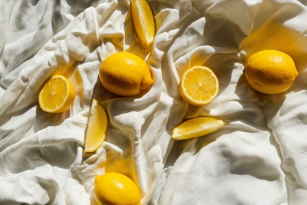 Lemon-themed fabric background with vibrant whole and sliced lemons. Bright yellow lemons contrast sharply against white fabric texture, creating a sunny, fresh still life aesthetic. Key elements: yellow lemons, white background, slices and whole fruit.