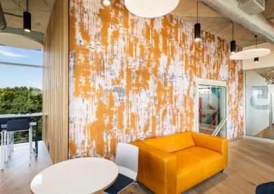 The wallpaper is textured with a blend of orange and white elements. It features an abstract, modern design that complements the bold orange couch. The pattern intricately combines various shades of orange with clean white accents, creating a dynamic yet cohesive look that adds depth to the office lounge space.