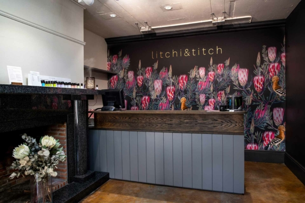 The wallpaper features a black background adorned with an intricate floral mural. The design includes vibrant pink and peach litchi flowers, lush green foliage, and subtle white accents that create a striking contrast against the dark backdrop. The detailed floral elements add a touch of elegance to the modern store interior.