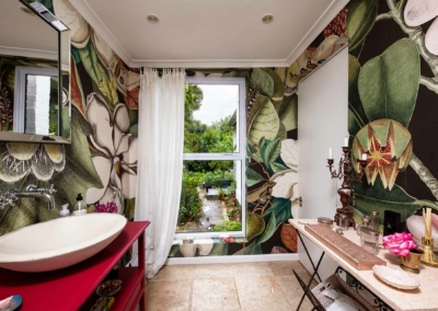 Our vibrant bathroom features a stunning large botanical wallpaper, showcasing lush green leaves, vivid tropical flowers in shades of pink, red, and orange, and intricate vines. This striking design sets a lively backdrop with its rich array of natural elements and vibrant colors.