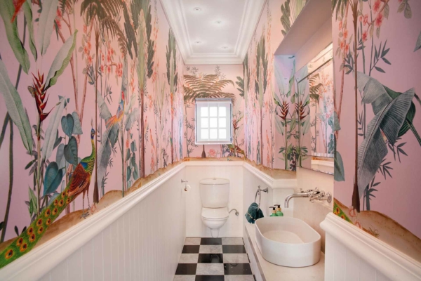 The wallpaper in this vibrant, tropical-themed bathroom features a lively array of illustrated plants and animals. Bright pink serves as the background, setting off intricate illustrations of peacocks with their dazzling feathers spread wide and playful monkeys swinging through lush greenery. The colorful foliage includes deep emerald leaves and exotic flowers in shades like coral, turquoise, and gold. Bold black patterns add depth to the design, making this wallpaper a striking focal point in any space.