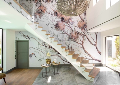 A nature-themed mural wallpaper features elements like lush green leaves, floral motifs, and delicate branches. Earthy tones of greens, browns, and touches of soft blues dominate the design. The artwork brings in a lively yet serene ambiance with organic shapes inspired by nature's elegance. Ideal for making a space feel fresh and vibrant.