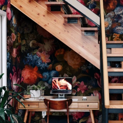 The vibrant floral wallpaper under the floating wooden staircase features elements like blooming roses, delicate peonies, and lush green leaves. The colors are rich and varied, including deep reds, soft pinks, bright yellows, and fresh greens. This lively pattern serves as a colorful backdrop for the cozy workspace setup.