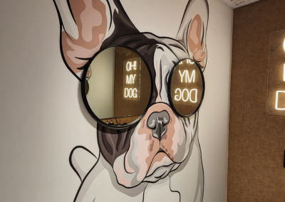 The wallpaper features a painted mural of a French Bulldog wearing large, reflective sunglasses with the text "OH MY DOG." The design includes a ceiling adorned with paw print patterns. Colors include various shades of gray for the bulldog, metallic hues for the sunglasses, and white for the text. The paw prints on the ceiling are black against a lighter background.