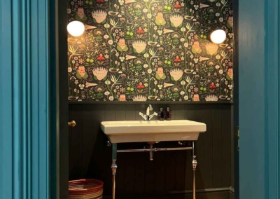 Dark floral wallpaper features rich, deep black background adorned with vibrant blooms in shades of burgundy, deep blue, and gold. Intricate green leaves and delicate vine tendrils weave through the flowers. Hints of ivory and mauve add contrast and dimension to the pattern.
