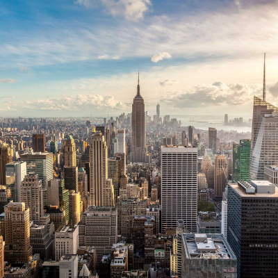 Aerial view wallpaper of a cityscape with tall skyscrapers under a partly cloudy sky. Highlighted by the iconic Empire State Building in the center. Features both modern and classic architecture. River visible in the distance. Dominant colors: gray, white, blue, and hints of green.