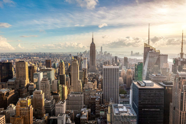 Aerial view wallpaper of a cityscape with tall skyscrapers under a partly cloudy sky. Highlighted by the iconic Empire State Building in the center. Features both modern and classic architecture. River visible in the distance. Dominant colors: gray, white, blue, and hints of green.