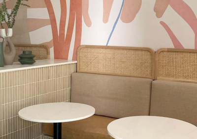 A cozy café interior features a wall mural in soft pastel colors. The wallpaper boasts delicate patterns with shades of mint green, blush pink, light lavender, and soft yellow. Intricate designs include floral motifs and geometric shapes that create a serene and inviting atmosphere. This patterned wall mural acts as a tranquil backdrop to the space's other design elements.