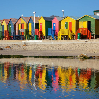 Colorful wooden beach huts in a neat row on sandy beach, painted red, yellow, green, blue. Vibrant reflections in calm water. Clear blue sky. Serene coastal vineyard setting.